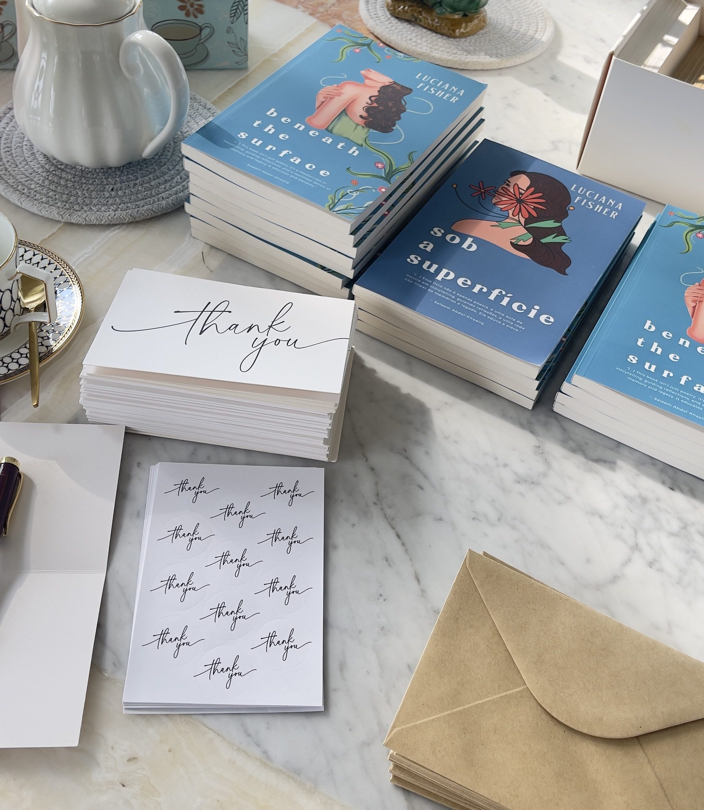 A table display featuring stacks of books by Luciana Pissarra, a teapot, a thank you card, and envelopes on a marble surface, accented by a potted plant.