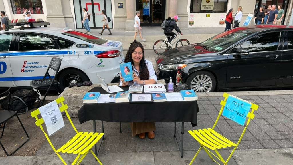 A woman at a table displaying books, with a yellow chair beside her that has a sign saying 'rest (and read)'. A police car is parked nearby, and people are walking by on the street.